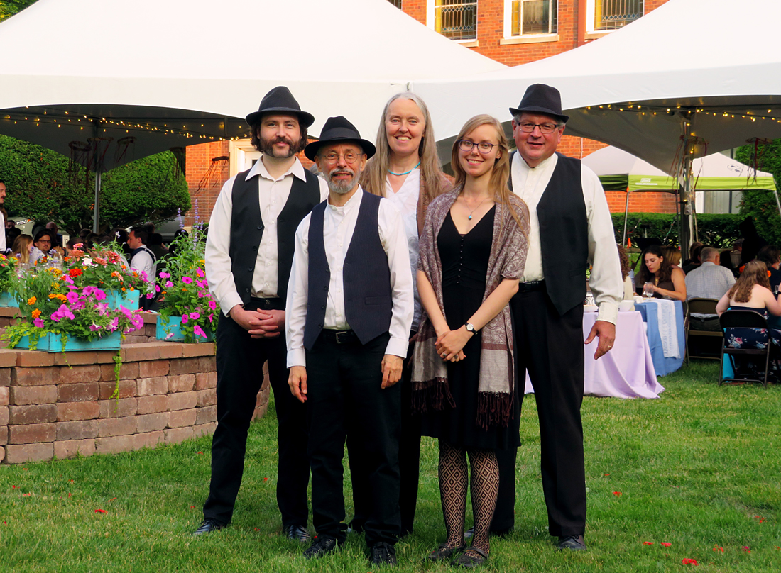 Photo of five musicians standing in front of white, peaked-top tents at an outside wedding